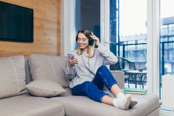 Young beautiful woman resting at home listening to fun music on headphones over the phone, online from the app and smiling - Foto, Bild