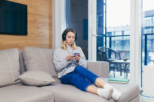 Young beautiful woman resting at home listening to audio podcast sitting on sofa, using headphones and smartphone, for online listening - Foto, Bild