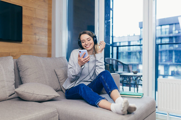 Young beautiful woman resting at home listening to fun music on headphones over the phone, online from the app and smiling - 写真・画像