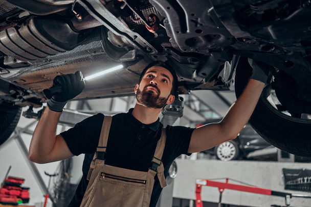Professional auto mechanic working on the undercarriage of a car. Diligence, attention, inspection, examination, annual checkup, safety, insurance, professionalism, vehicle concept. Bottom view. - Photo, image