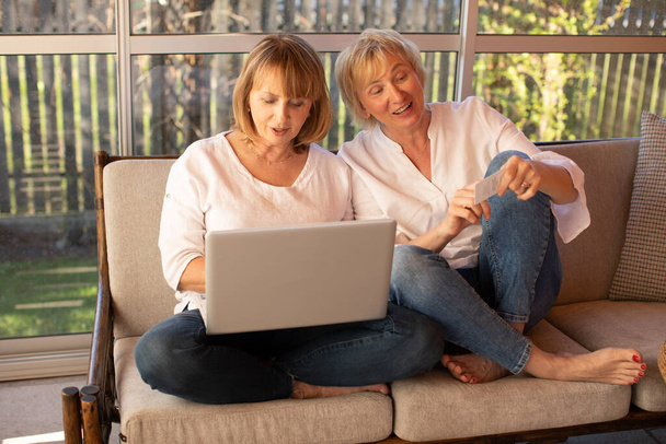 Две 55-летние женщины в повседневной одежде используют ноутбук, чтобы совершать покупки в Интернете, удобно сидя дома на диване - Фото, изображение