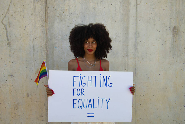 Young, beautiful African-American woman claiming equality of people with a banner and a gay pride flag. - Photo, Image