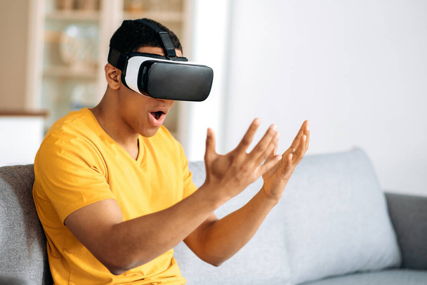Un tipo hispano atónito y moderno probando gafas VR. Un joven usa gafas de realidad virtual, mira excitado a sus manos, gestos con las manos, sentado en casa en el sofá de la sala de estar - Foto, imagen