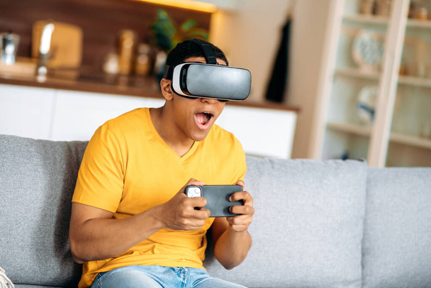Heureux jeune homme hispanique assis sur un canapé à la maison en vêtements décontractés, utilise des lunettes VR, appareil de réalité virtuelle, dans un monde numérique, tient smartphone, jouer dans le monde virtuel - Photo, image