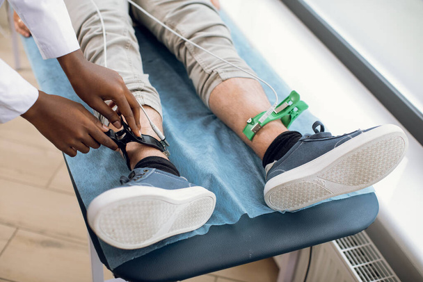 ECG concept. Close up cropped top view of male patient legs, lying on the couch in hospital, while female afro american doctor fixing sensor electrodes for electrocardiogram recording. - Photo, Image