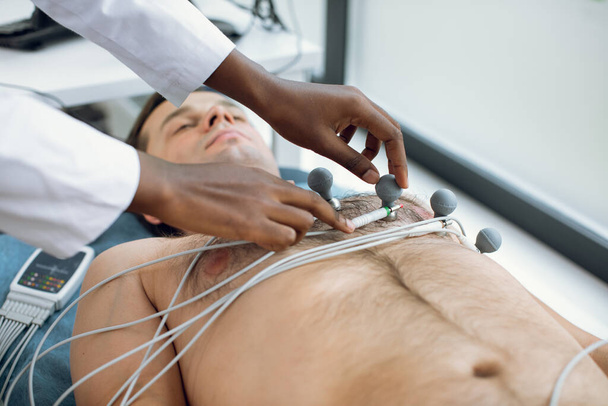 Cropped image of female african american doctor or nurse sticking holter and vacuum electrodes on young male patients chest in hospital. ECG and heart health concept. Annual health check. - Foto, immagini