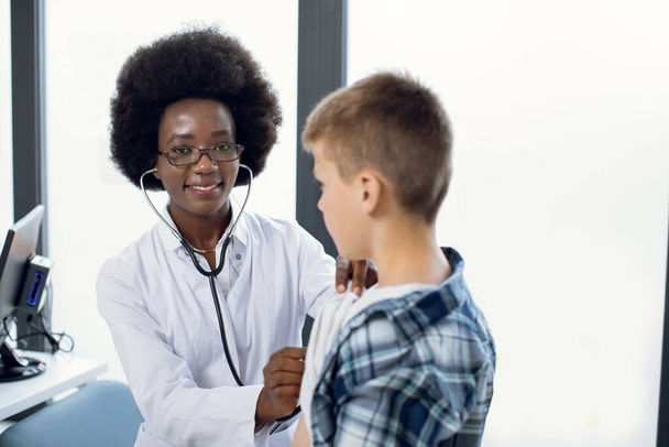 Young smiling pleasant African female pediatrician with stethoscope auscultating kid boy patient. Black physician, checks heartbeat and lungs breathing of kid, making checkup before vaccination. - Foto, imagen