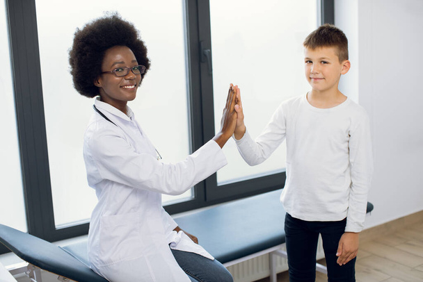 Young pretty likable medical worker, Afro-American lady doctor gp pediatrician, giving her cute child boy patient a high five after successful annual medical check up in modern clinic. - Фото, изображение