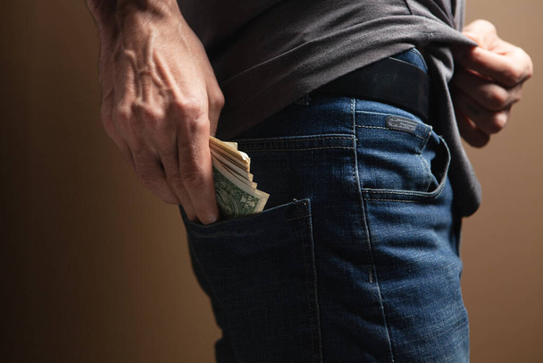 Ο άνθρωπος βάζει λεφτά στην πίσω τσέπη του σε καφέ φόντο. - Φωτογραφία, εικόνα