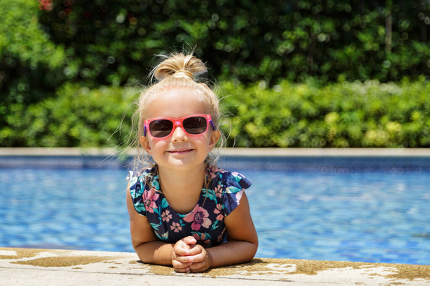 Happy little caucasian toddler girl with sunglasses smiling joyfully and enjoying summer vacations time in swimming pool in resort hotel. Kid with blonde hair wearing swimwear during summer holidays  - 写真・画像