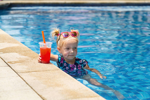 Feliz niña pequeña caucásica con gafas de sol sonriendo alegremente y disfrutando de las vacaciones de verano en la piscina en el hotel resort. Niño con el pelo rubio usando trajes de baño durante las vacaciones de verano  - Foto, imagen