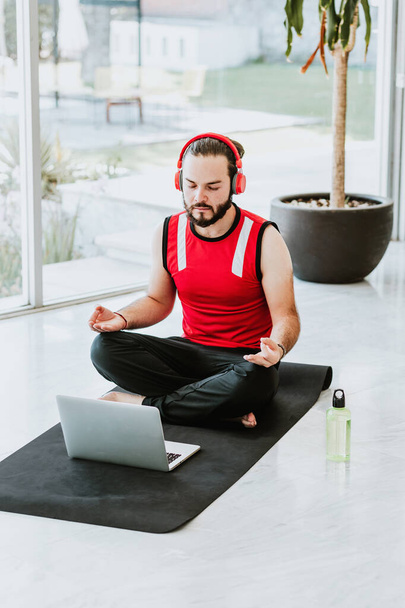 赤いヘッドフォンを持つラテン人はノートパソコンでヨガセッションでリラックスするためにガイド付き瞑想を聞いて - 写真・画像