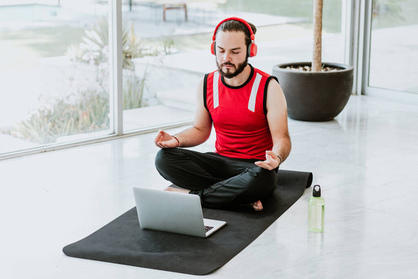 Latinský muž s červenými sluchátky poslouchá řízenou meditaci k relaxaci v józe sezení na notebooku - Fotografie, Obrázek