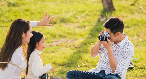 Šťastné asijské mladé rodiny otec, matka a dítě baví a těší venkovní společně sedí na travnaté party s focením fotografií retro fotoaparát piknik v zahradním parku na slunný den - Fotografie, Obrázek