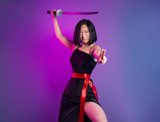 スレンダーなアジア系の女性で、刀を手にしてネオンを背景に侍をイメージした黒いドレスを着ている。 - 写真・画像
