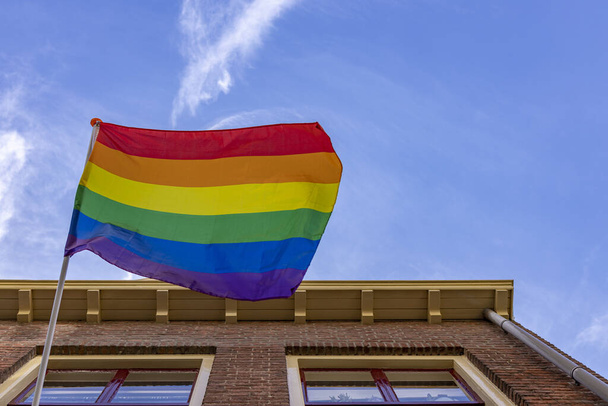 Rainbow colored LGBTQ+ flag contrasted strongly against a clear blue sky waving in a strong wind seen from below hanging from a Dutch exterior facade - Zdjęcie, obraz