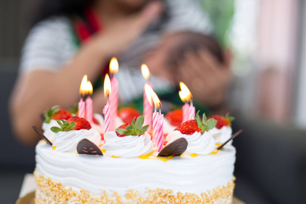 A strawberry birthday cake with lit candles and a caring mother holding a newborn baby in the background - Фото, изображение