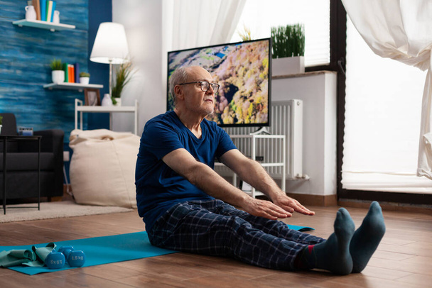 Pensionato concentrato che indossa abbigliamento sportivo seduto sul tappetino yoga che allunga il muscolo delle gambe - Foto, immagini