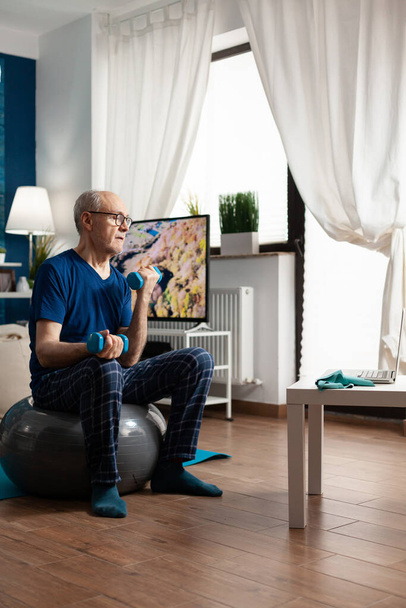 Senior man training body resistance doing arms exercise sitting on swiss ball in living room - Photo, Image