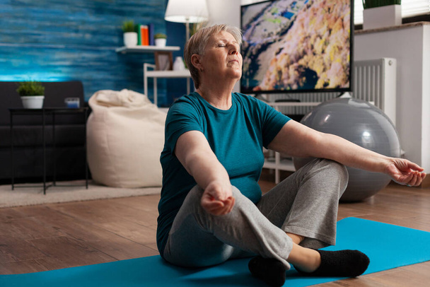 Mujer mayor sentada cómoda en posición de loto en esterilla de yoga con los ojos cerrados - Foto, Imagen
