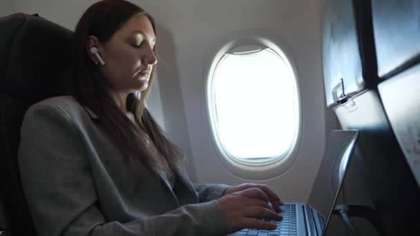 Brünette Frau im Anzug und mit drahtlosem Kopfhörer im Ohr tippt im Flugzeug Text auf einen Laptop - Filmmaterial, Video