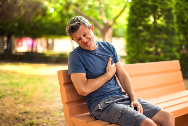 Человек с болью в сердце на улице. Мужчина чувствует боль в груди, сидя на скамейке в парке. Здравоохранение и медицина  - Фото, изображение