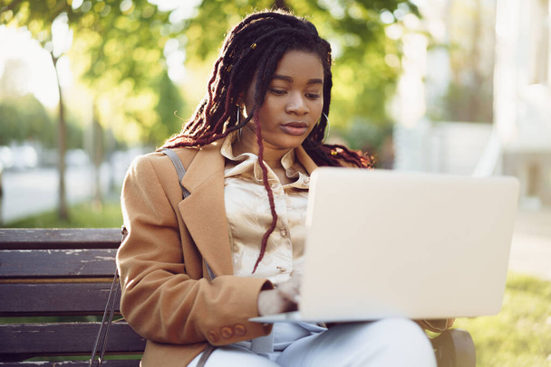 Африканская американка-фрилансер сидит на скамейке на улице и пользуется ноутбуком - Фото, изображение
