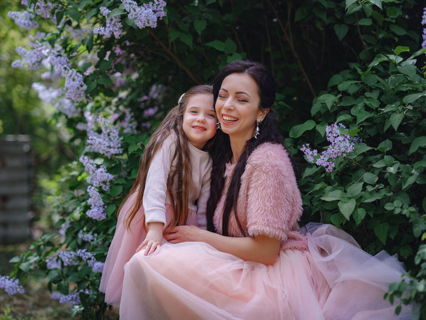 γυναίκα με κόρη Σε όμορφα ροζ φορέματα διασκεδάζοντας σε εξωτερικούς χώρους την άνοιξη λιλά κήπο. Η μαμά και το παιδί χαμογελούν και αγκαλιάζονται. Οικογενειακές διακοπές και συντροφικότητα. Ευτυχισμένη Ημέρα της Μητέρας! - Φωτογραφία, εικόνα