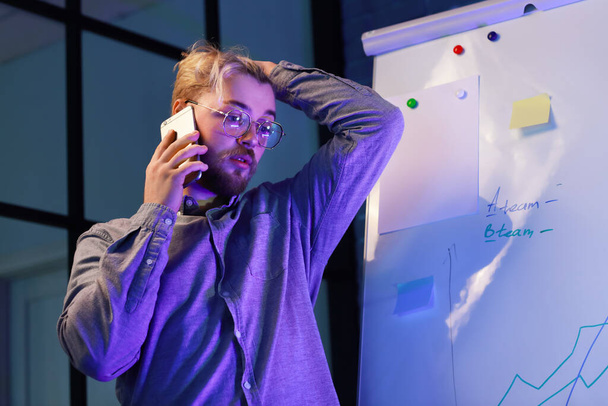 Väsynyt nuori mies puhuu matkapuhelimella toimistossa yöllä - Valokuva, kuva