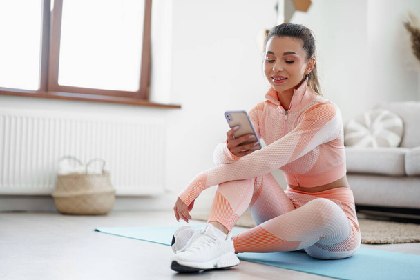 Beautiful young woman in sportswear sitting indoors and looking at smartphone before workout - Fotoğraf, Görsel