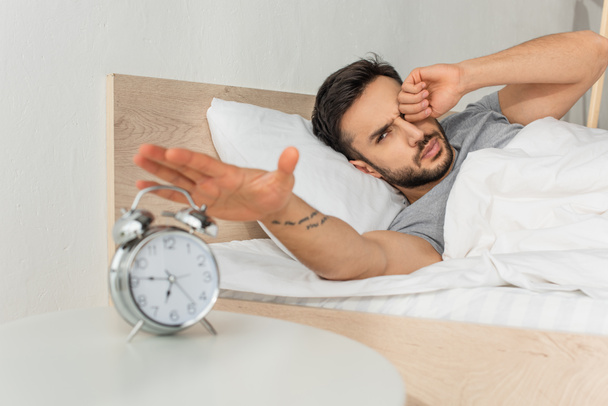 Young man pulling hand to blurred alarm clock while waking up  - Foto, afbeelding