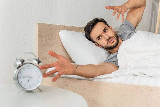 Homem irritado olhando para despertador desfocado na cama  - Foto, Imagem
