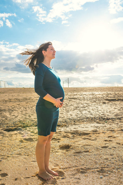 Pregnant woman in blue dress is standing on the sand and smiling, her hair blows. Calm and happy pregnancy lifestyle. - Photo, Image