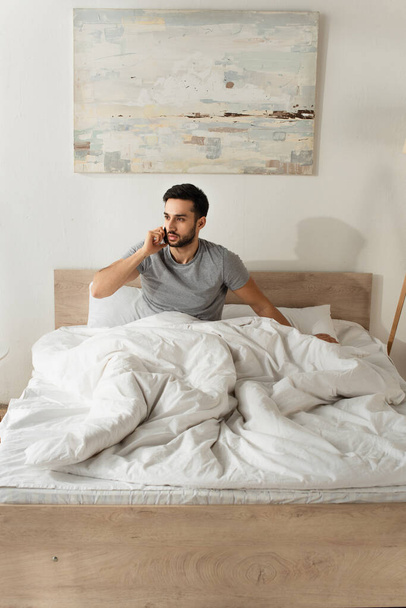 Bearded man sitting on bed and talking on smartphone  - Foto, imagen