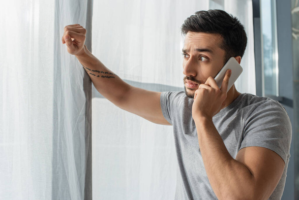 Hämmästynyt mies puhuu matkapuhelimella ja katselee ikkunaa kotona  - Valokuva, kuva