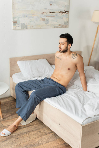 Мужчина без рубашки в штанах и тапочках сидит на кровати  - Фото, изображение