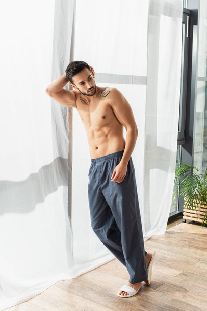 Shirtless man in slippers and pants standing near window  - Foto, imagen
