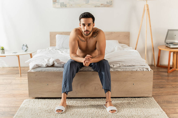 Mladý muž bez košile sedí na posteli a dívá se do kamery  - Fotografie, Obrázek