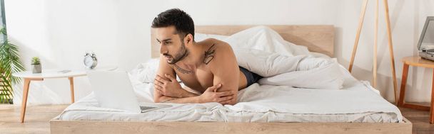 Shirtless freelancer looking at laptop on bed in morning, banner  - Фото, изображение