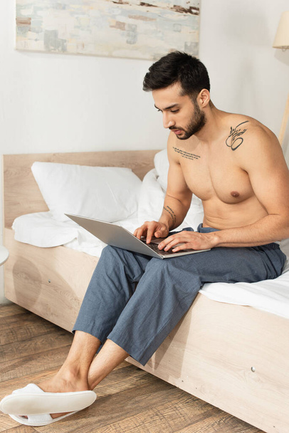 Ing nélküli férfi papucsban és nadrágban laptoppal az ágyon  - Fotó, kép