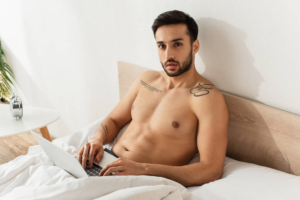 Shirtless freelancer using laptop on bed near alarm clock on bedside table  - Fotó, kép