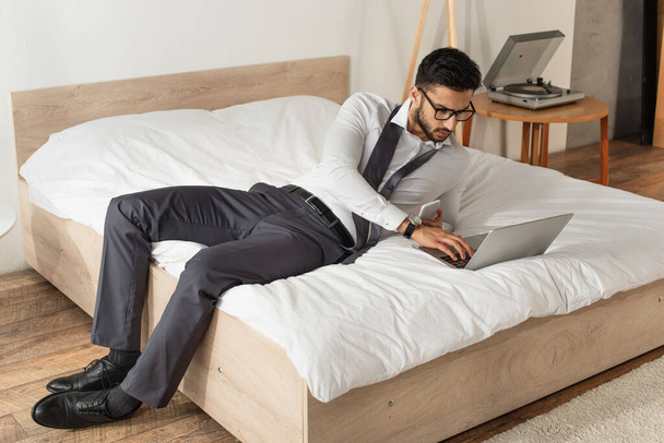 Homme d'affaires avec smartphone utilisant un ordinateur portable sur le lit à la maison  - Photo, image