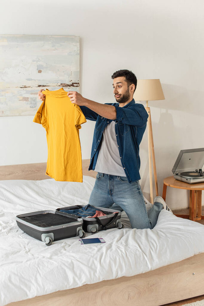 Aggressiver Mann sieht T-Shirt neben Koffer und Reisepass im Schlafzimmer  - Foto, Bild