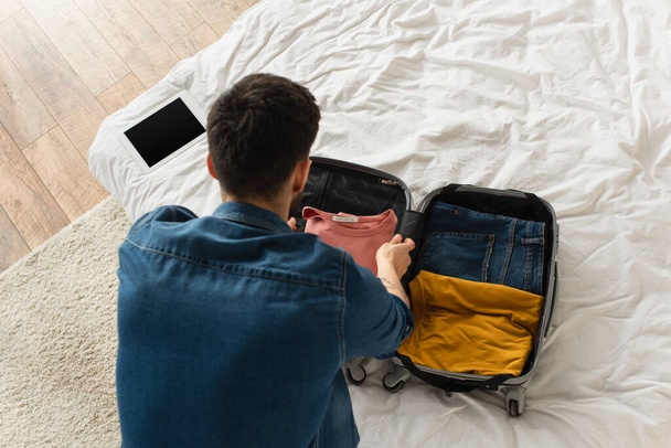 Overhead view of man putting clothes in suitcase near digital tablet on bed  - Foto, immagini