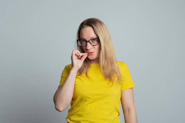 秘密を隠せ。大人の女性の肖像画では、キーの上に口を閉じているかのようにジップジェスチャーを示すメガネ,秘密を保つために約束,カジュアルな黄色のtシャツを着て,グレーの背景 - 写真・画像