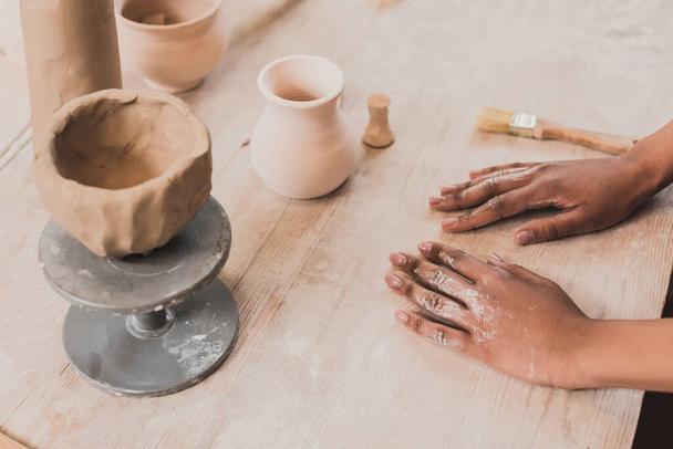 высокий угол зрения на глиняный горшок ручной работы на столе рядом с руками молодой африканской американки в керамике - Фото, изображение