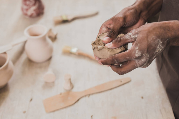 vista parziale di giovane uomo afroamericano scultura pezzo di argilla con le mani vicino al tavolo con attrezzature in ceramica  - Foto, immagini