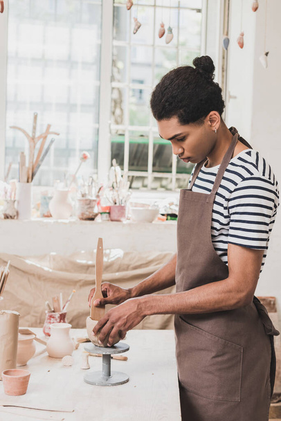 serio joven afroamericano hombre esculpir olla de arcilla con espátula en la mesa con equipo en cerámica  - Foto, imagen