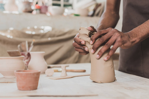 vista parziale di giovane uomo afroamericano che lavora con pezzo di argilla sul tavolo con attrezzature in ceramica  - Foto, immagini