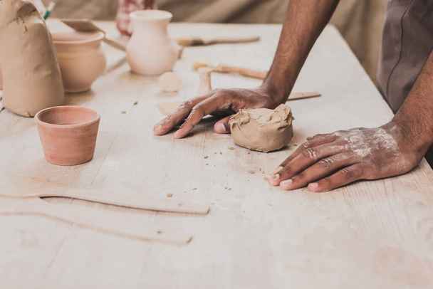 vista parziale di giovane uomo afroamericano con mani vicino pezzo di argilla sul tavolo con attrezzature in ceramica - Foto, immagini